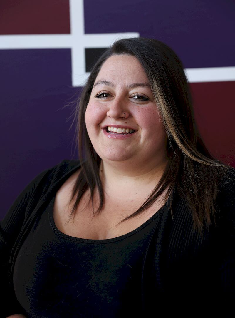 A smiling woman with long dark hair and a nose ring is wearing a black top. She is standing in front of a purple wall with white and brown geometric patterns.