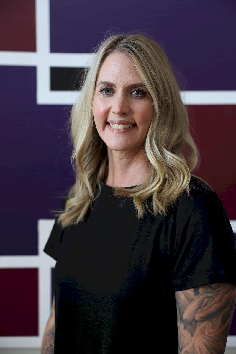 A person with long blonde hair smiles while standing in front of a geometric background with purple, red, and white shapes. They are wearing a black shirt and have tattoos on their arms.