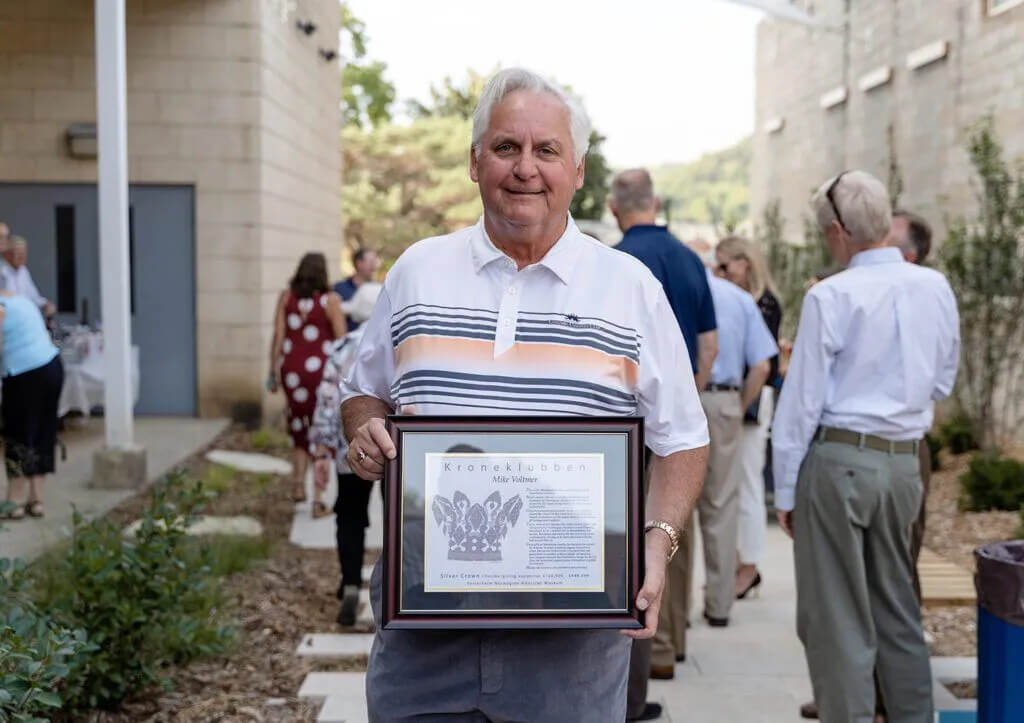 An older man stands outdoors holding a framed certificate. He's wearing a white and striped polo shirt. Several people in casual attire are gathered in the background, outside a building. Trees and plants are visible in the scene.