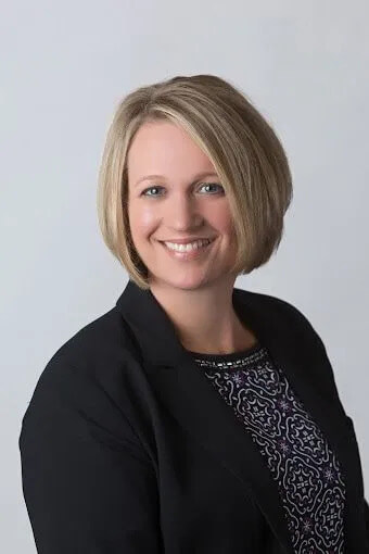 A person with short blonde hair is smiling at the camera. They are wearing a black blazer over a patterned top. The background is plain and light-colored.