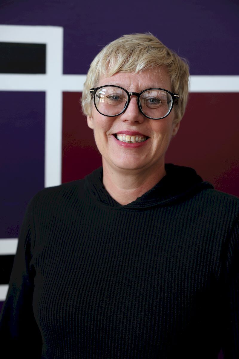 A person with short blonde hair and glasses smiles while standing in front of a geometric background with white, black, and maroon squares. They are wearing a black top.