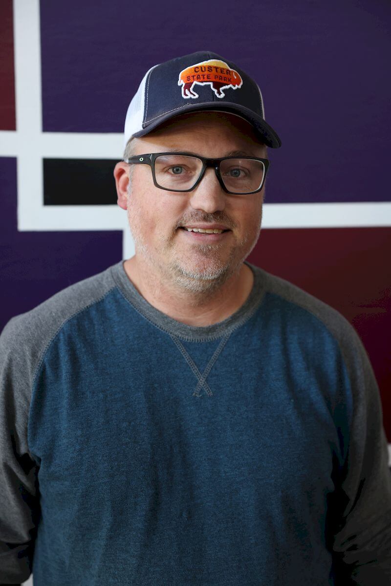 A man wearing glasses, a baseball cap with a buffalo emblem, and a blue shirt stands in front of a geometric background with purple and white colors.