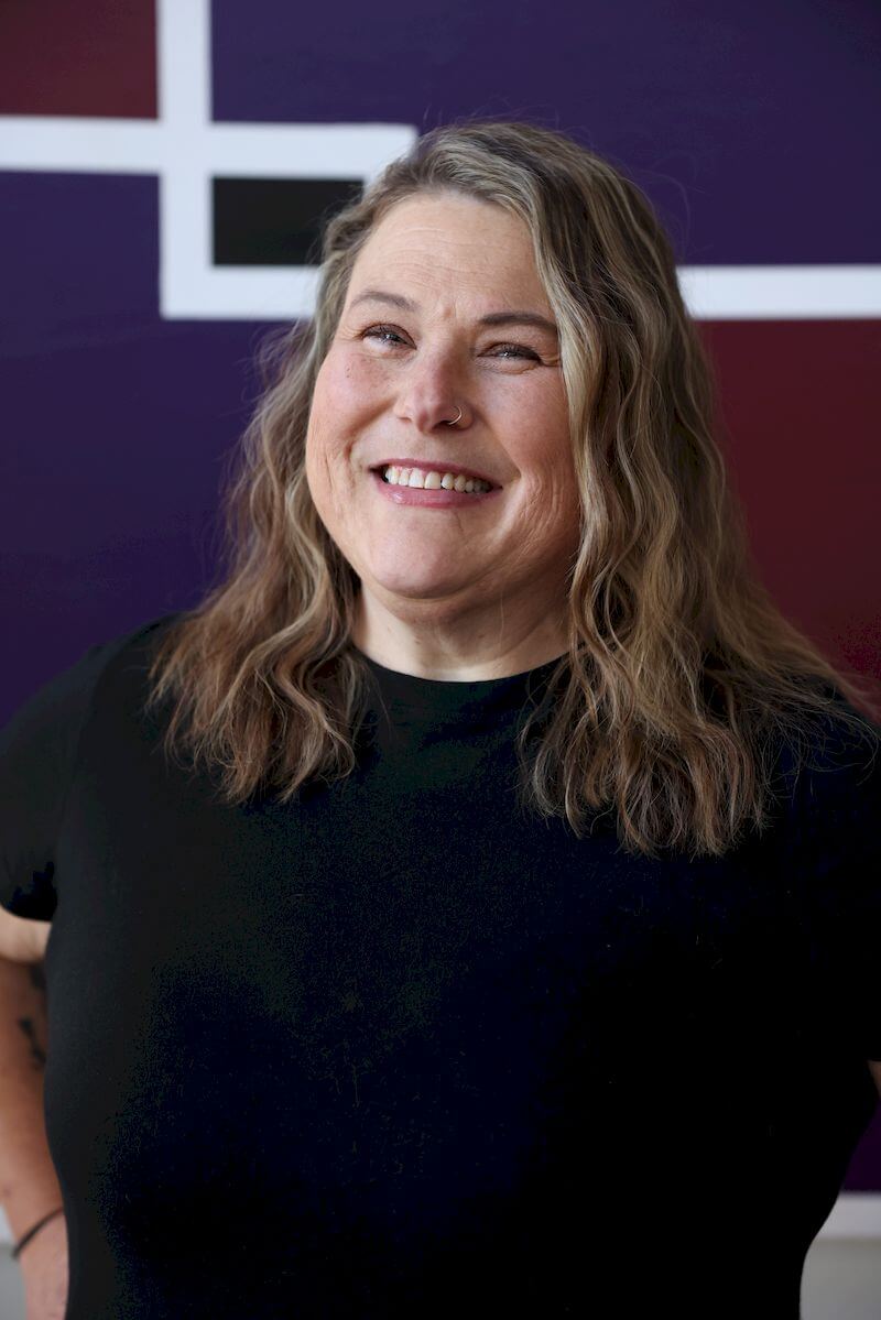 A person with long wavy hair, wearing a black shirt, is smiling in front of a purple and maroon geometric background.