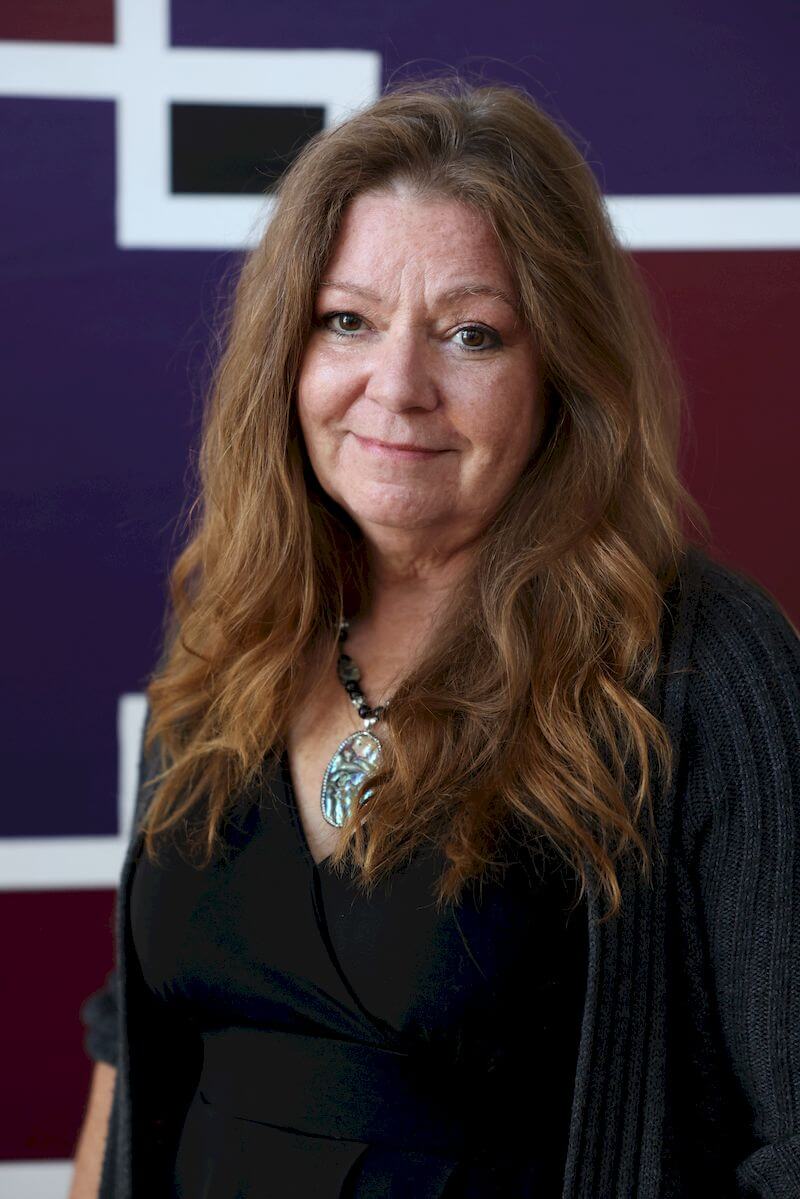 A woman with long, wavy hair wearing a black top and a gray cardigan stands in front of a purple and white geometric background. She is smiling slightly and wearing a large, decorative pendant necklace.