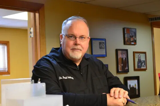A man with short gray hair and glasses is leaning on a counter, wearing a black shirt with embroidered text. Framed photos and certificates are visible on the wall behind him.